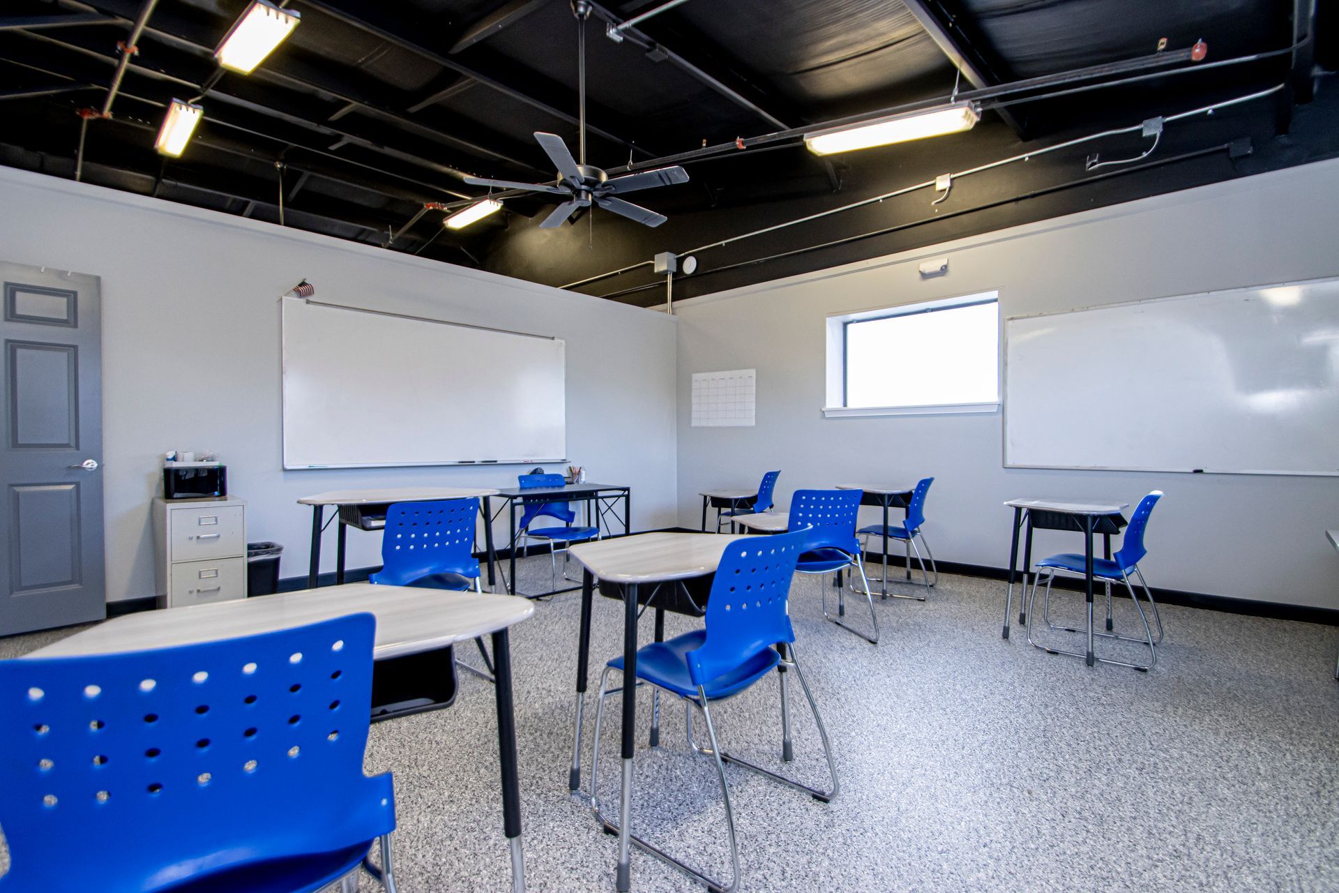 A classroom with white walls, blue chairs, desks, two whiteboards, and a dark ceiling with exposed lighting.