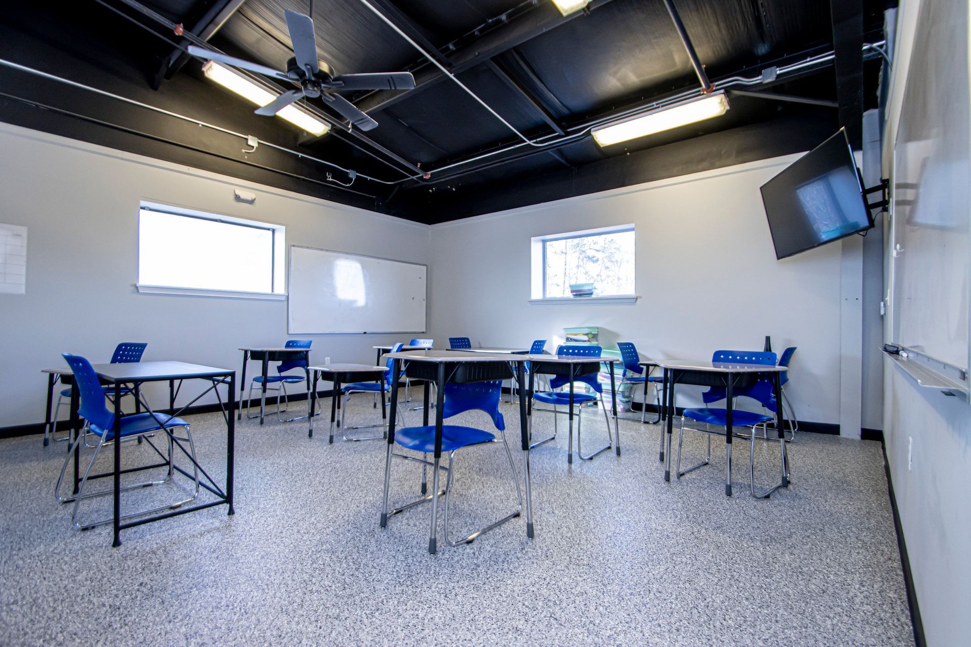 An empty classroom featuring white walls, a black ceiling, several desks with blue chairs, and whiteboards.