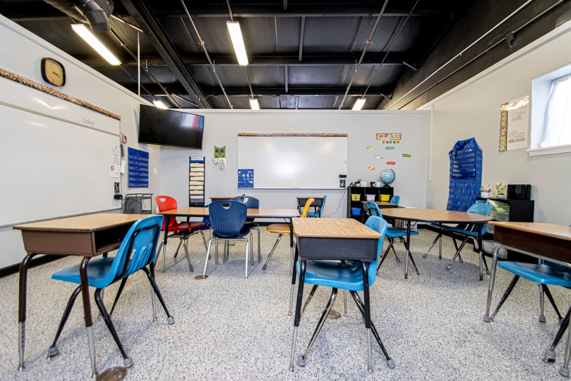 A bright classroom with white walls, a black ceiling, several desks with blue chairs, and whiteboards.