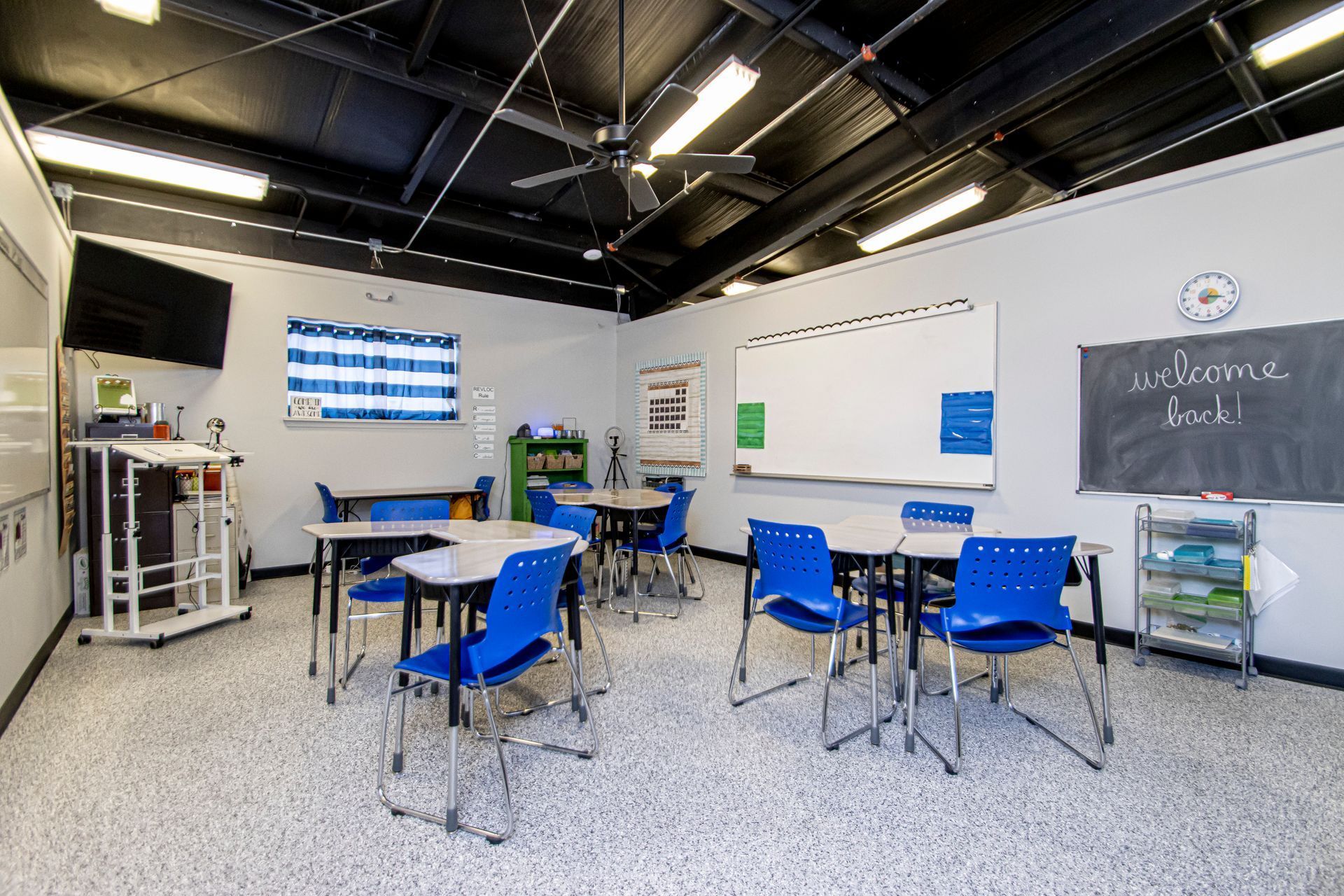 A brightly lit classroom with several student desks, blue chairs, a whiteboard, a chalkboard, and a small TV mounted.