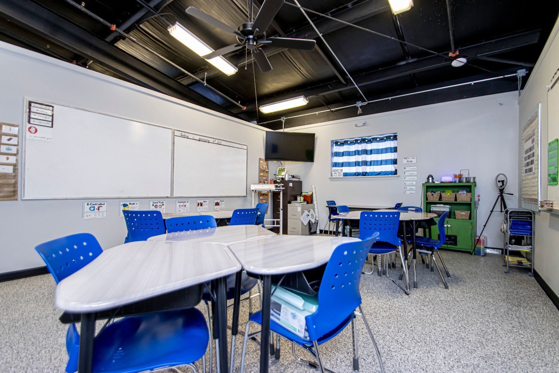 A classroom with grey walls, a black ceiling, two whiteboards, several blue chairs, and grouped trapezoidal desks.