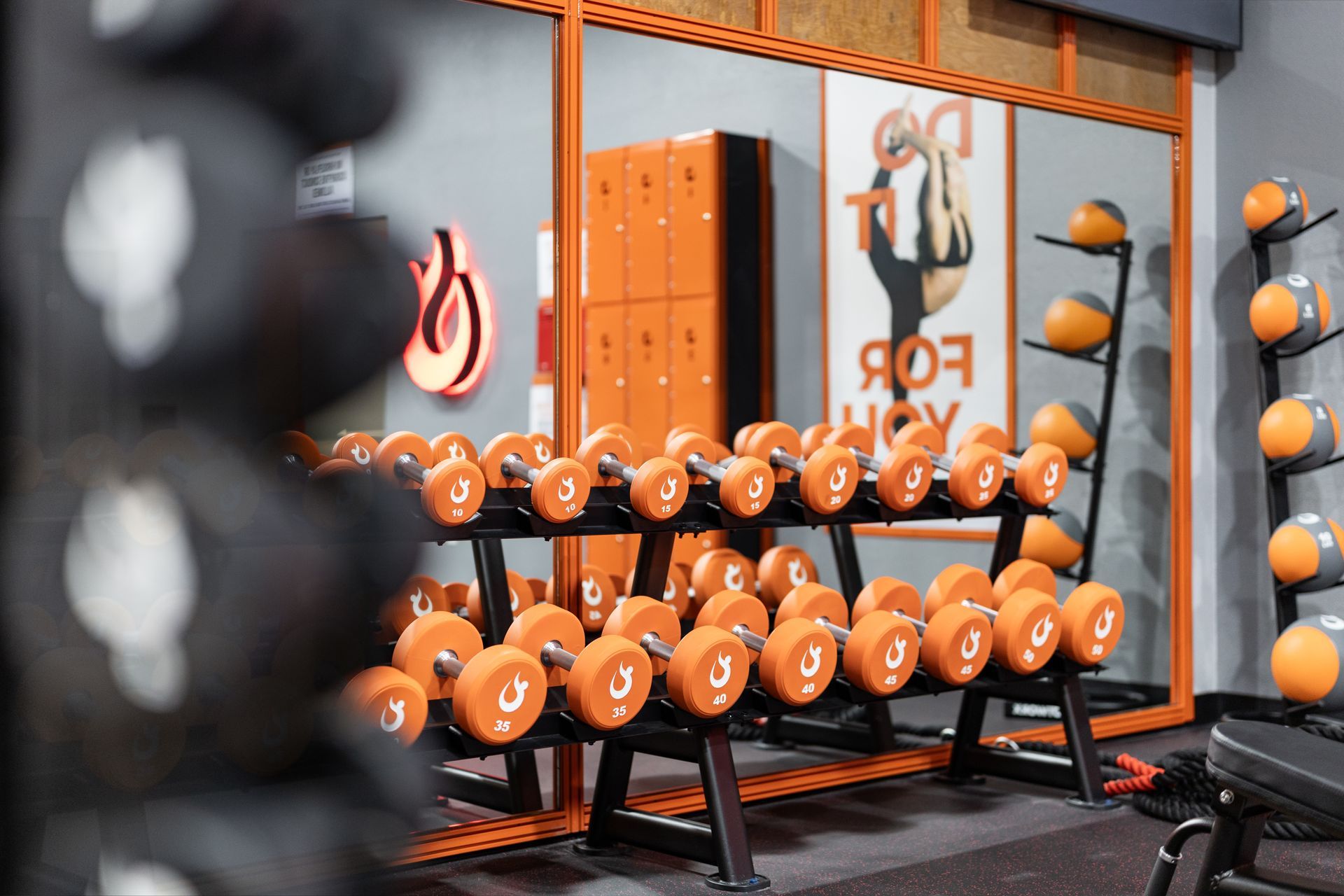 A gym interior with rows of orange dumbbells on racks, medicine balls, and orange lockers reflected in a large mirror.