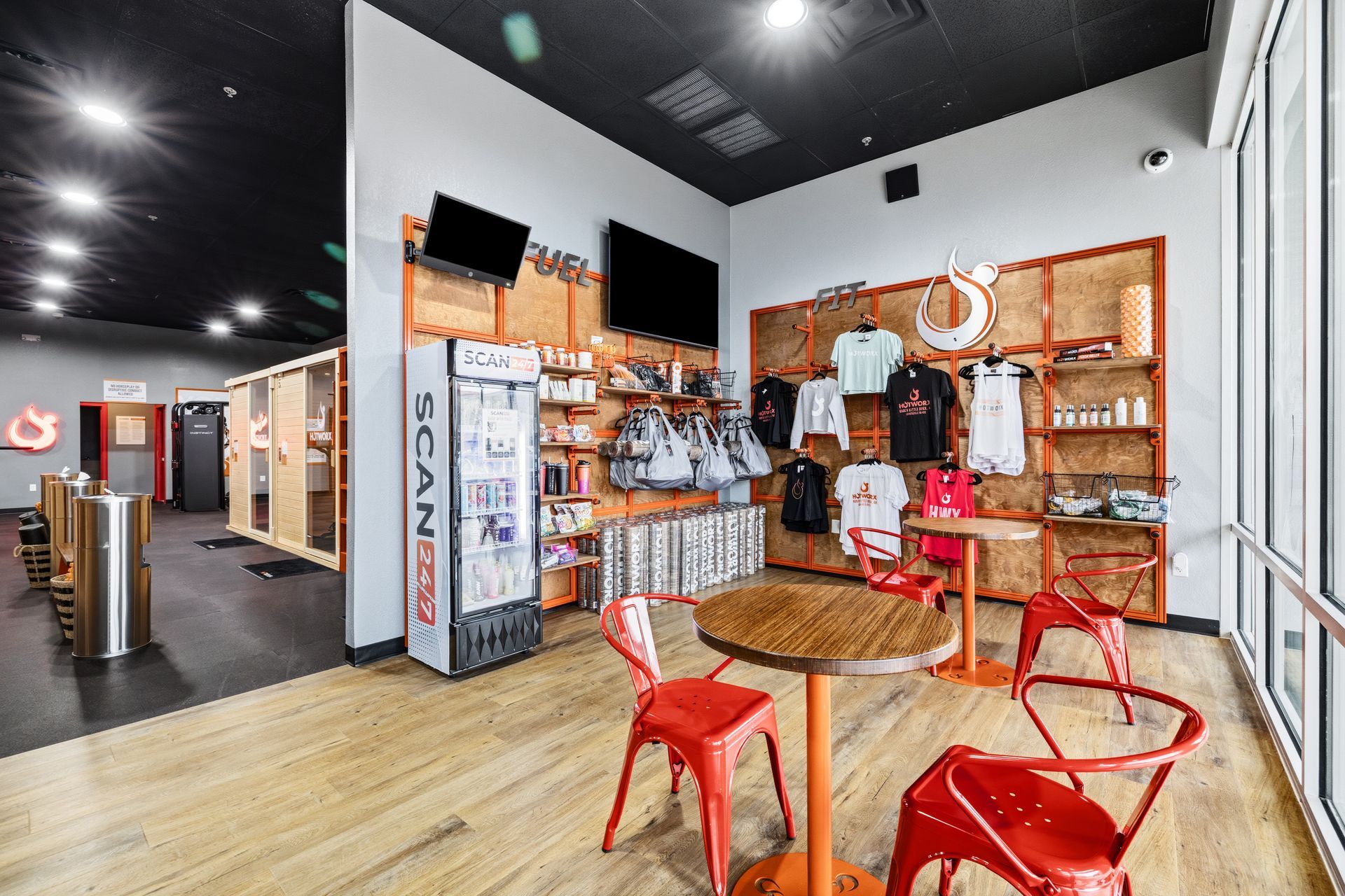 A fitness center lobby with wood floors, red metal chairs, a branded refrigerator, and apparel on display shelves.