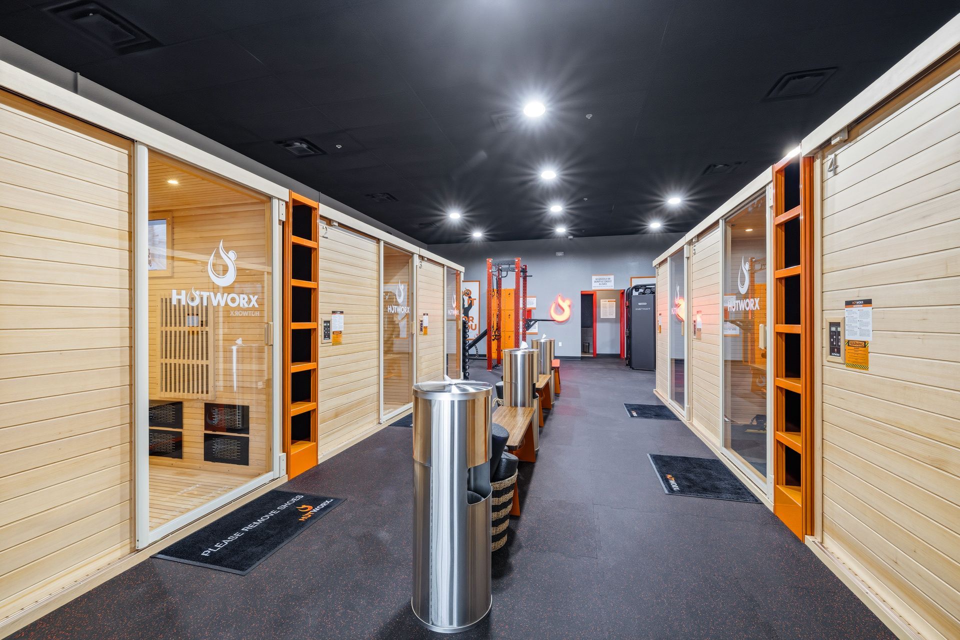 A gym interior with a central aisle between two rows of wooden Hotworx sauna workout pods under a dark ceiling.