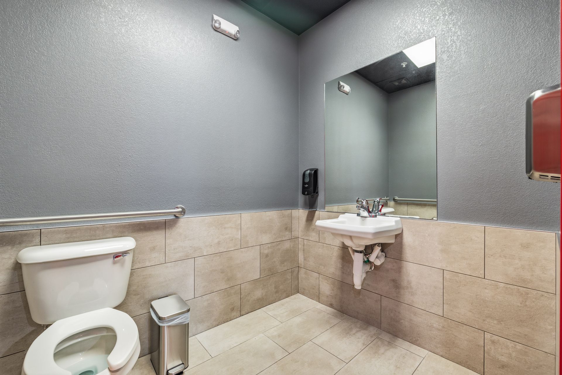 An accessible restroom with a white toilet, sink with a faucet, large mirror, and gray and tan tiled walls.