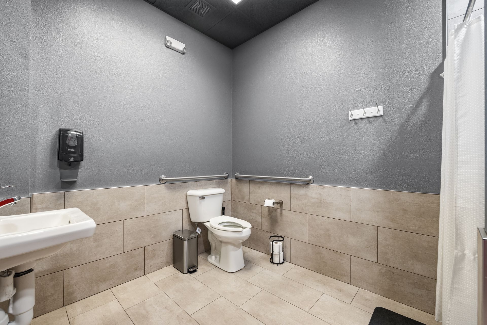 A spacious, accessible restroom featuring a white toilet with grab bars, a sink, and a shower curtain against gray walls.