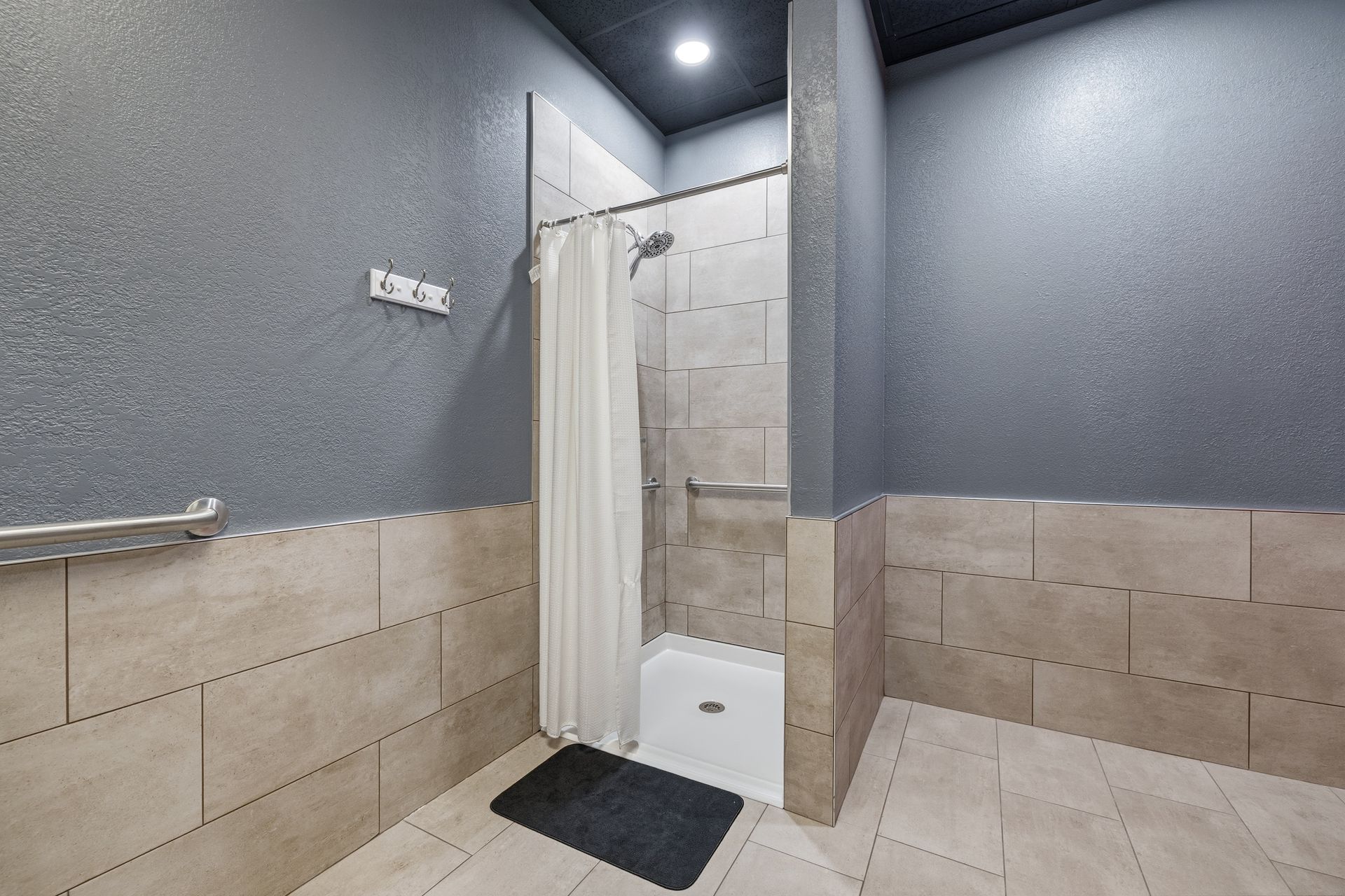 A handicap-accessible shower stall with beige tile, white shower curtain, and silver grab bars against dark gray walls.