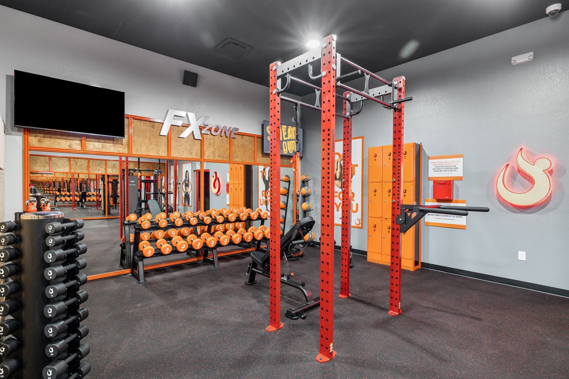 An indoor gym showing a red squat rack, a dumbbell rack with weights, and exercise benches on a dark rubber floor.