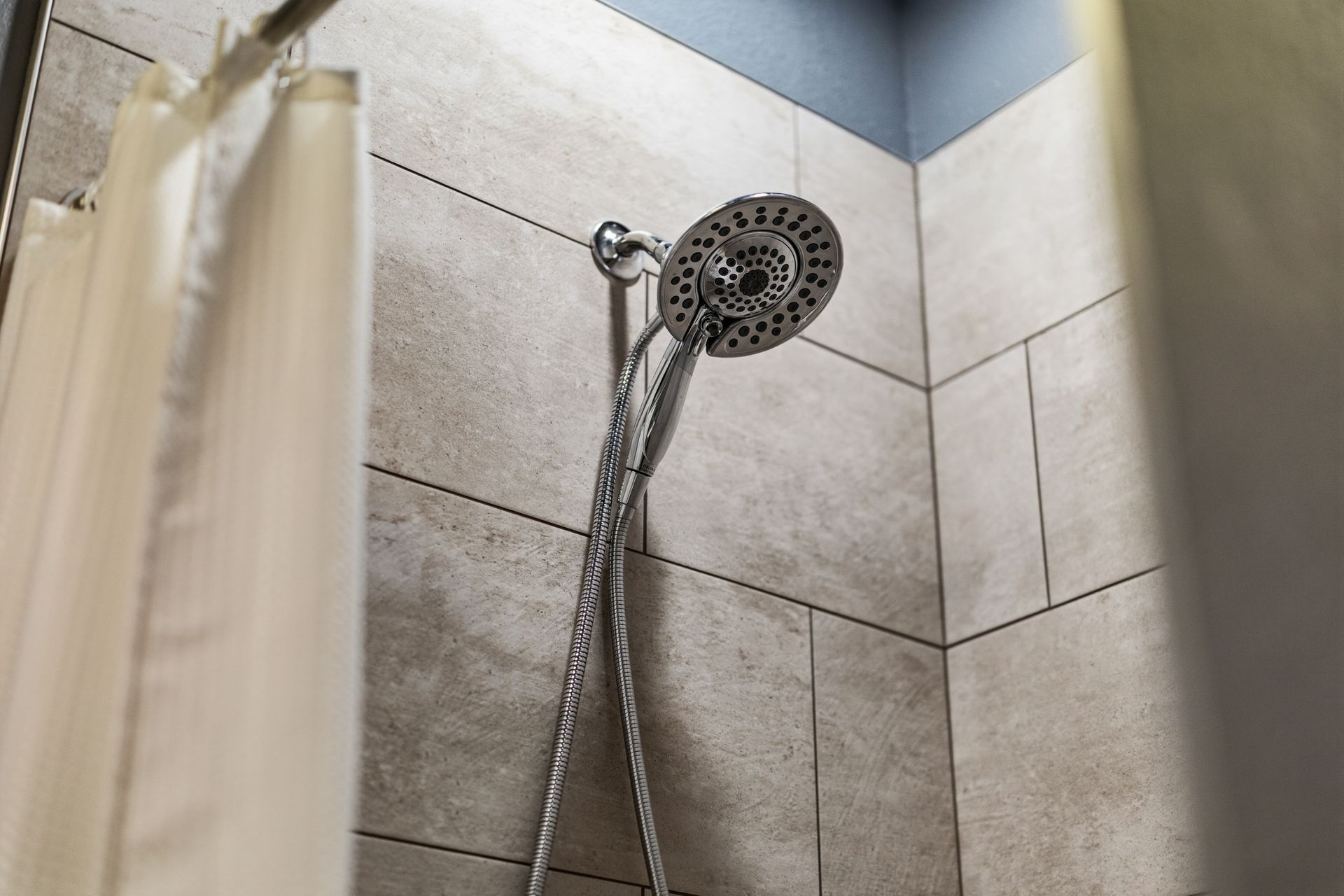 A handheld chrome shower head mounted on a light-colored, tiled bathroom wall next to a white shower curtain.