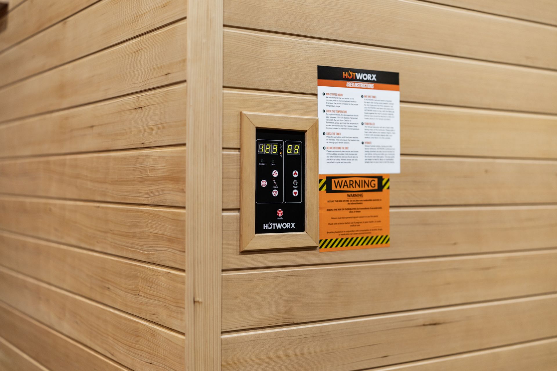 A wooden sauna wall with a digital control panel, operation instructions, and a warning label attached.