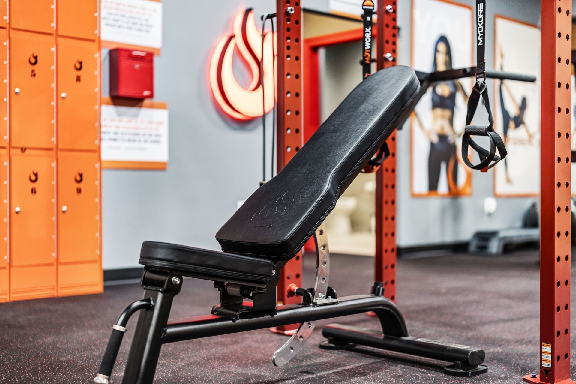 An adjustable black weight bench centered in a gym with orange lockers and fitness equipment in the background.