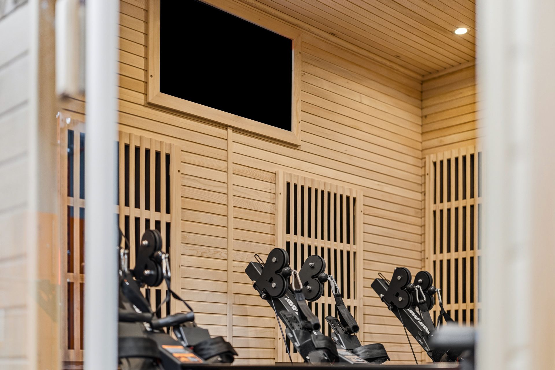 Rowing machines lined up against a wooden-paneled wall inside a gym, viewed through a doorway.
