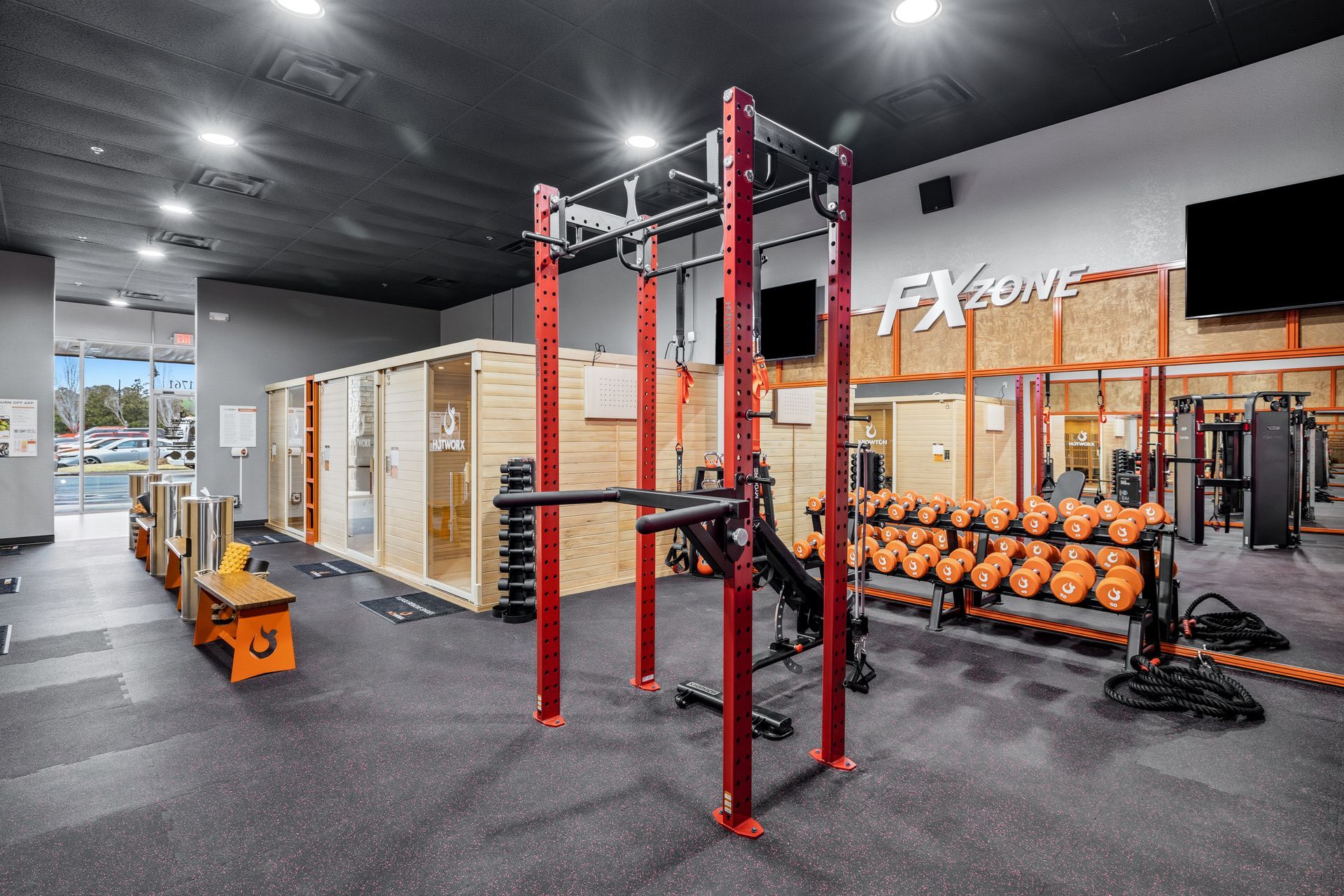 A brightly lit fitness center featuring a red power rack, a rack of dumbbells, and wooden sauna enclosures.