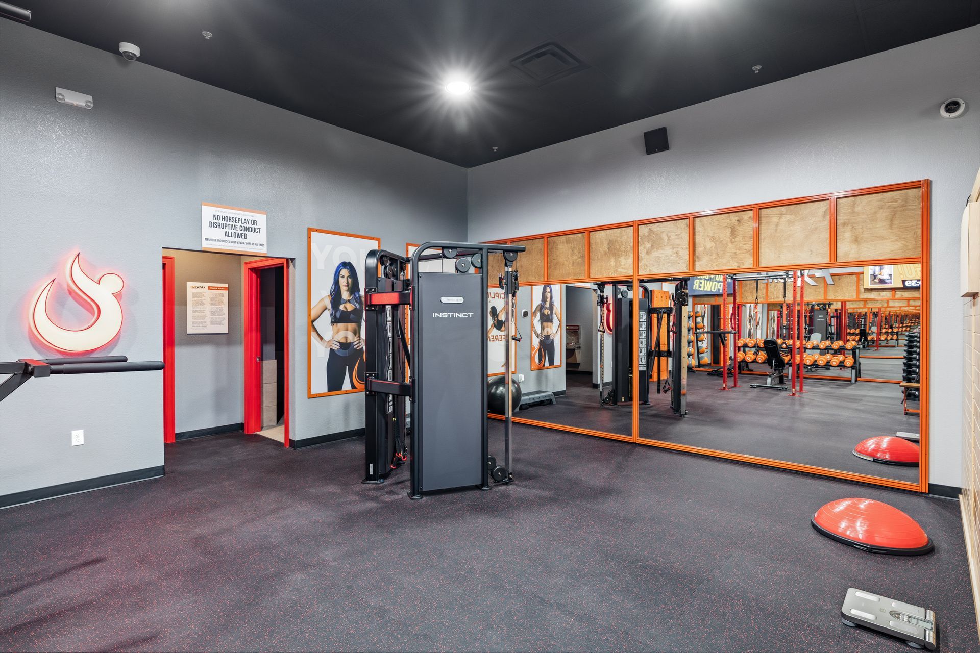 A modern gym interior with a black ceiling, gray walls, a cable machine, large wall mirrors, and an orange balance ball.