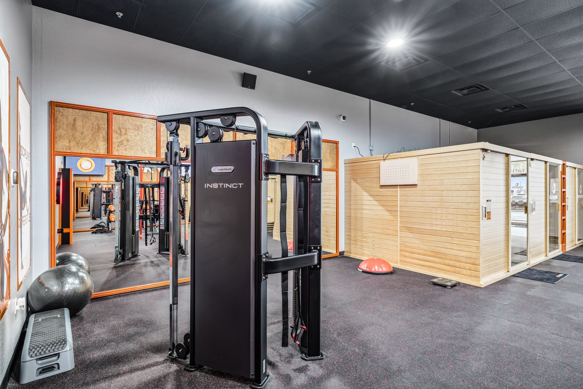 A gym interior with a black Technogym cable machine, a wooden sauna structure, and exercise balls on a dark floor.
