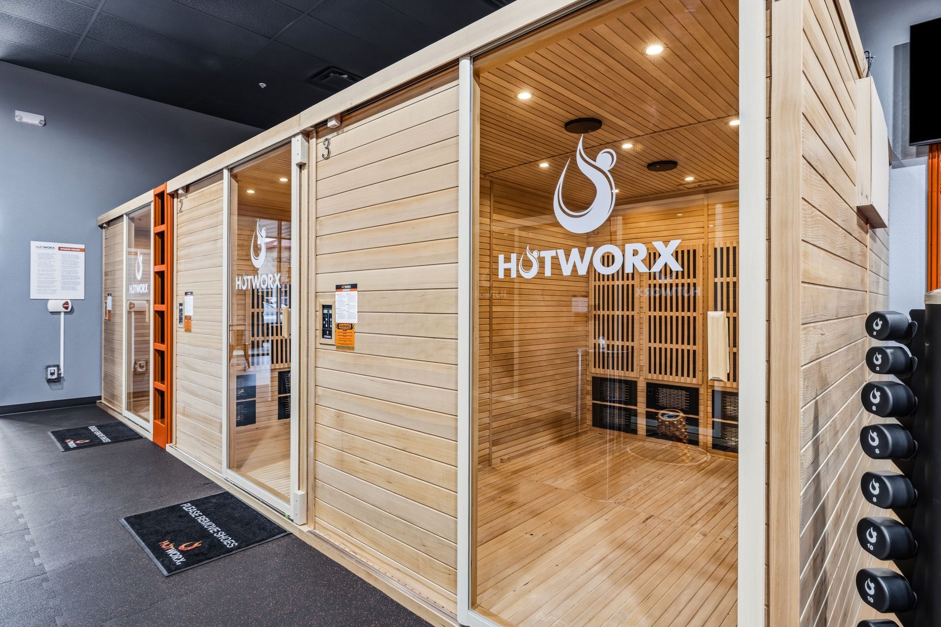 A row of wooden HOTWORX infrared sauna workout pods in a gym setting with a rack of dumbbells in the foreground.