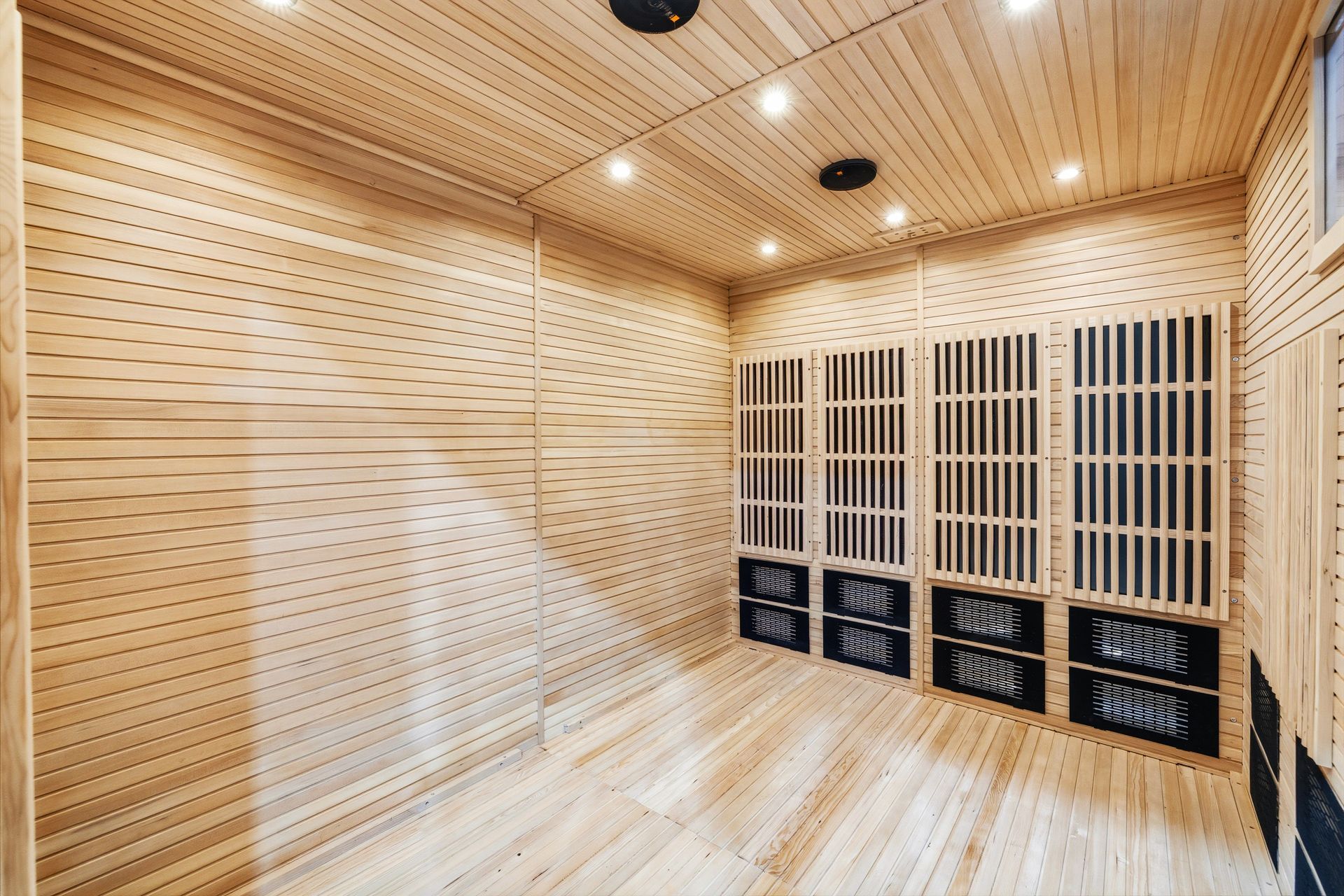 An empty, light-wood sauna featuring slat-covered wall heating panels and recessed ceiling lights.
