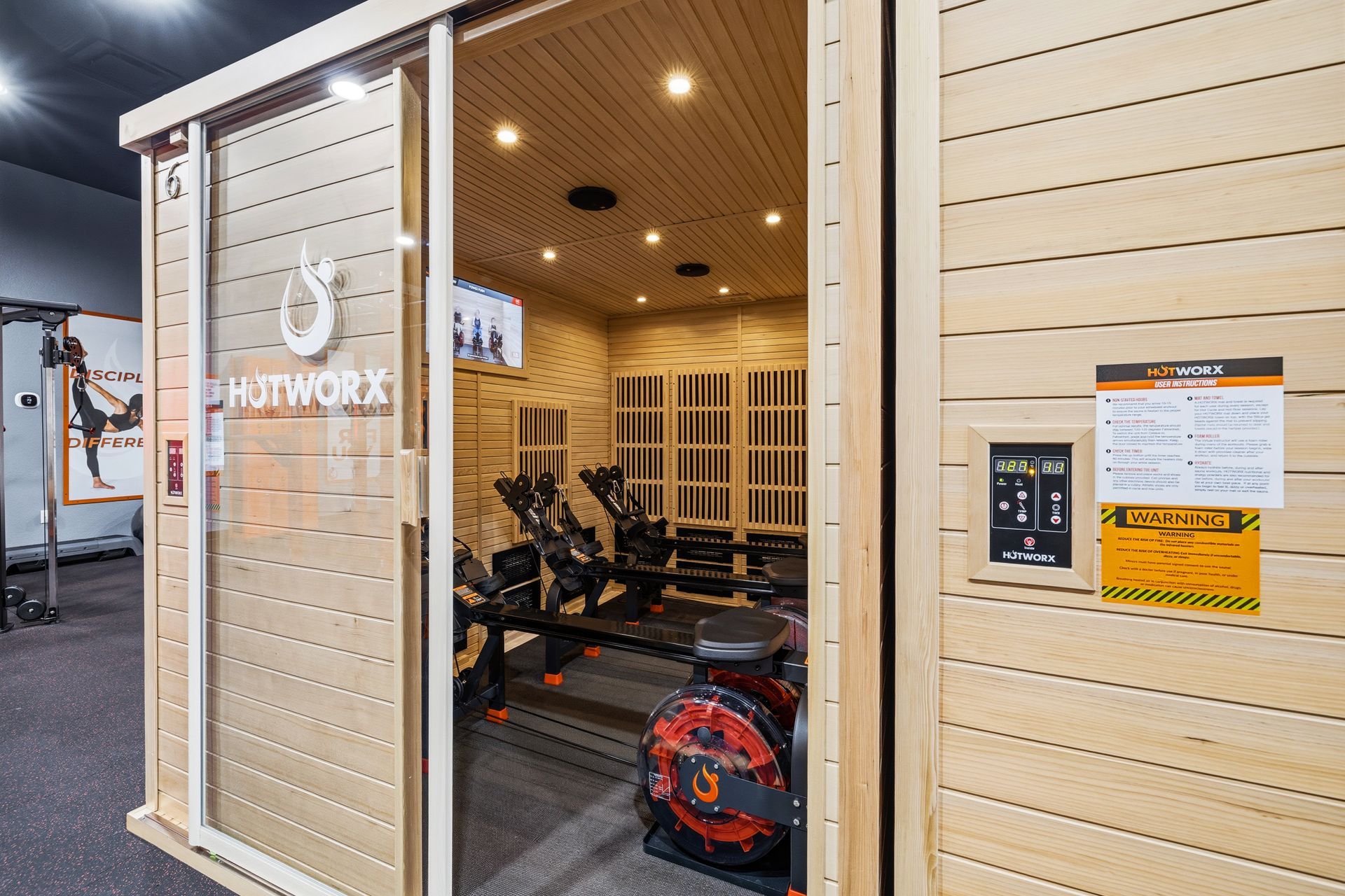 A HOTWORX sauna room at a fitness center featuring interior rowing machines, a television, and exterior control panels.