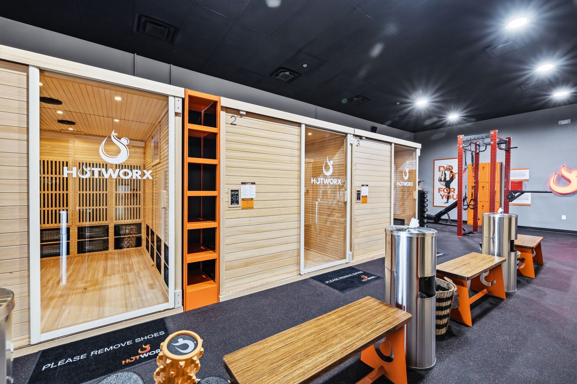 A Hotworx studio interior with wooden saunas, orange shelving, benches, and fitness equipment in a modern gym setting.