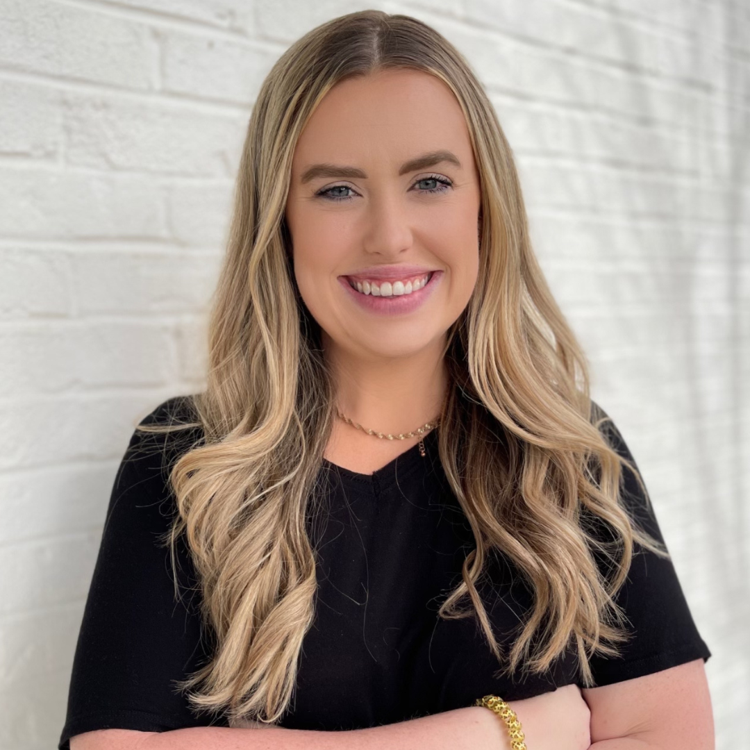 A smiling person with long blonde hair wearing a black top and gold chain necklace, standing against a white brick wall.