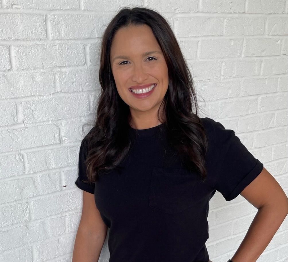 A smiling person with long dark hair wearing a black t-shirt, standing against a white painted brick wall.
