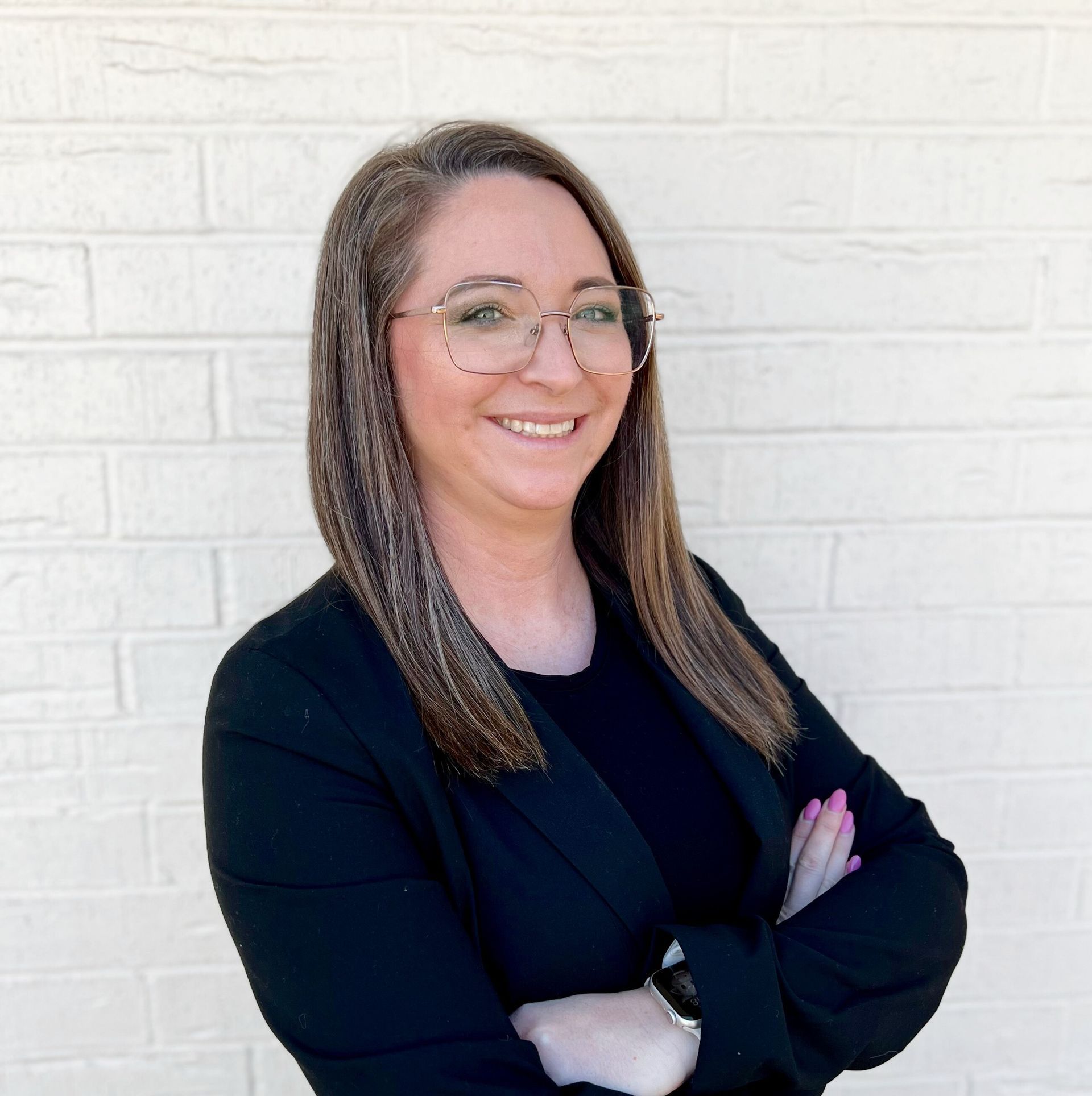 A person with long brown hair and glasses smiling with arms crossed, wearing a black blazer against a white brick wall.