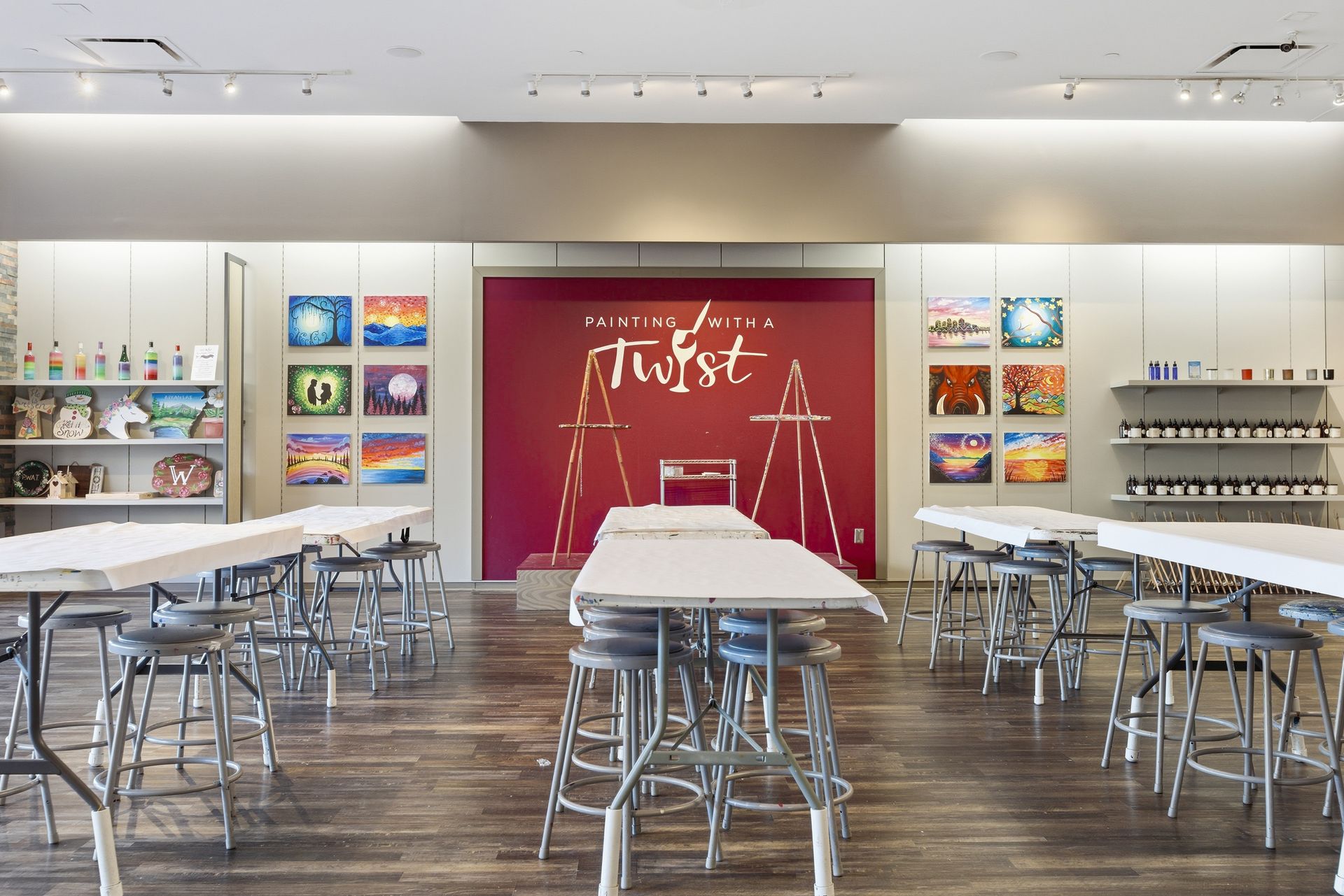 An interior view of a Painting with a Twist art studio, featuring rows of white tables, bar stools, and art displays.
