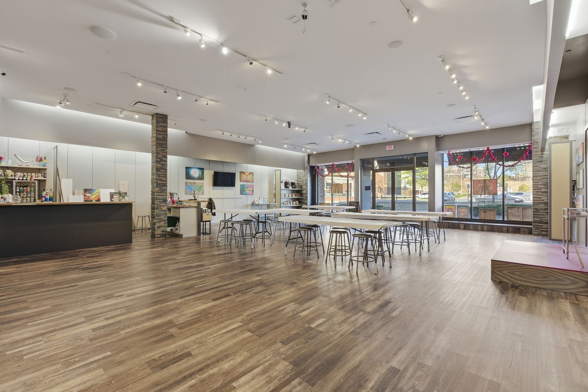 A spacious, brightly lit studio with wooden floors, rows of white tables and stools, and a large front window.