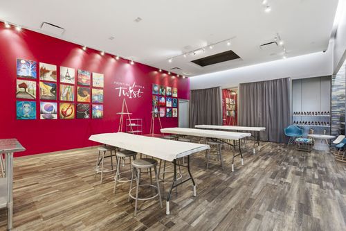 A painting studio with tables, stools, and a red wall featuring a grid of framed artwork and the words 