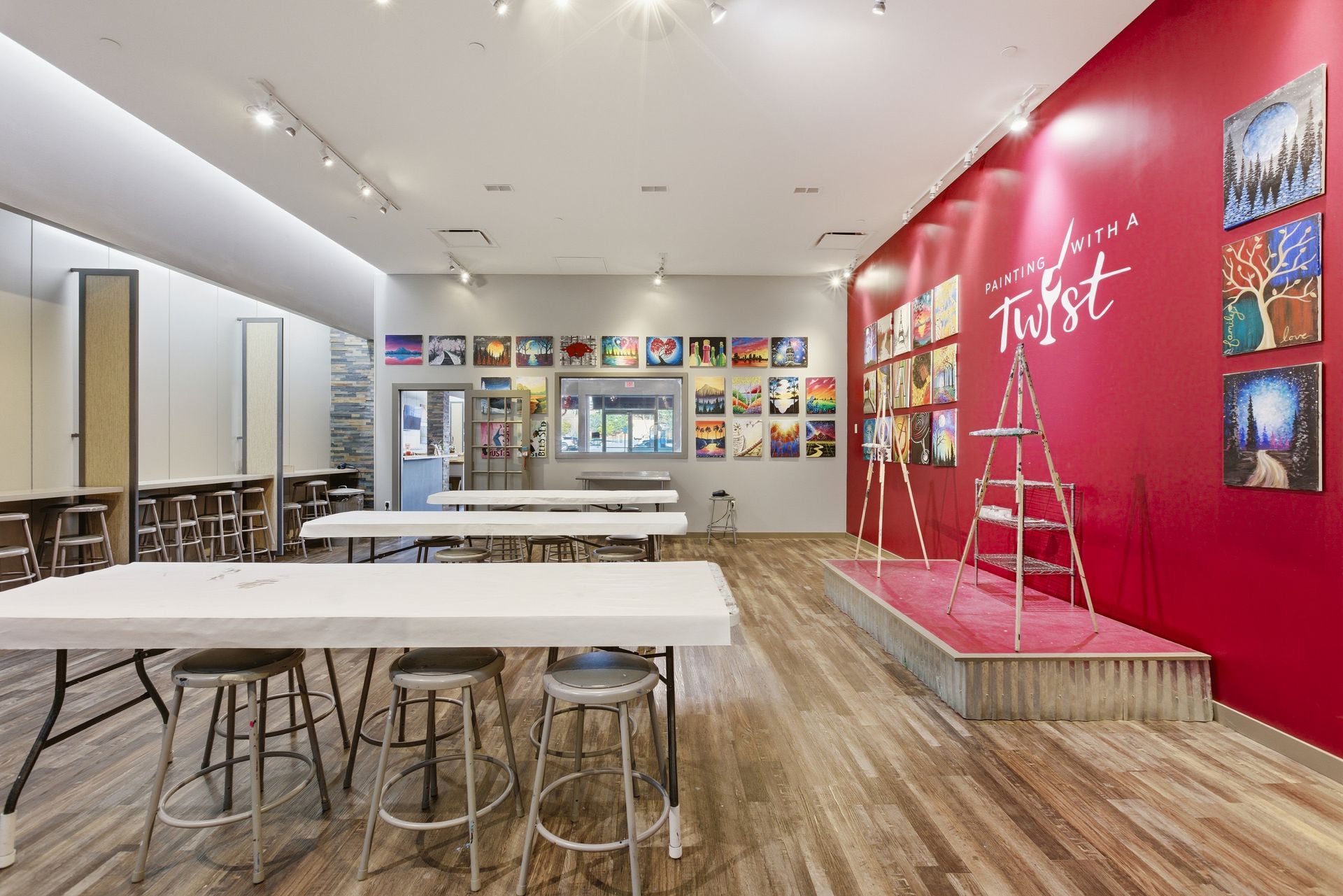 Empty painting studio with white tables, stools, a stage, and colorful paintings displayed on a bright red accent wall.