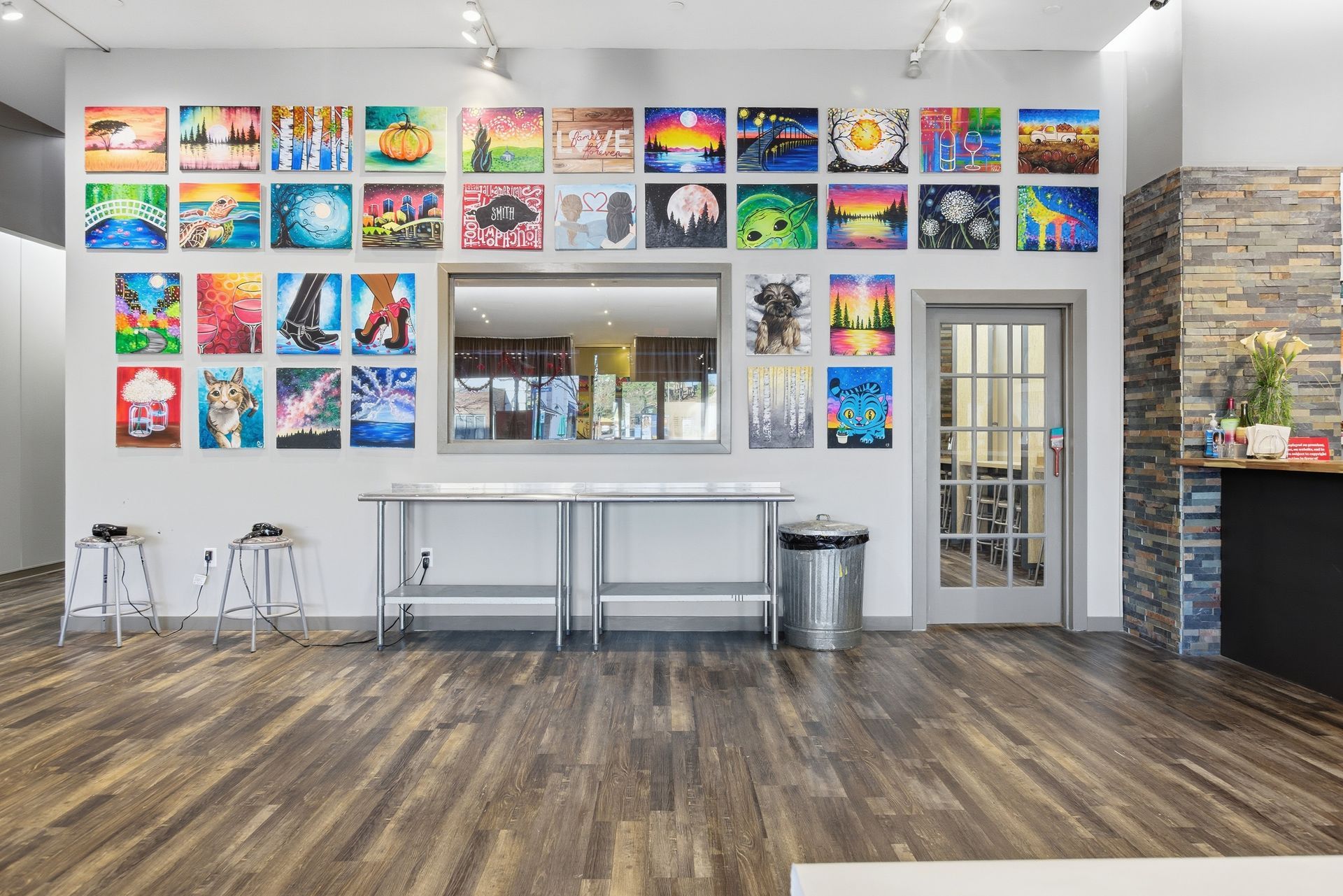 Art studio wall featuring rows of colorful paintings, a large central window, a stainless-steel table, and a trash bin.