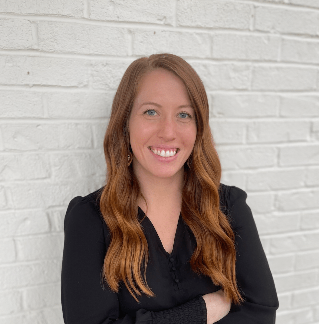 A person with long wavy auburn hair smiles, wearing a black top, posing against a white brick wall.