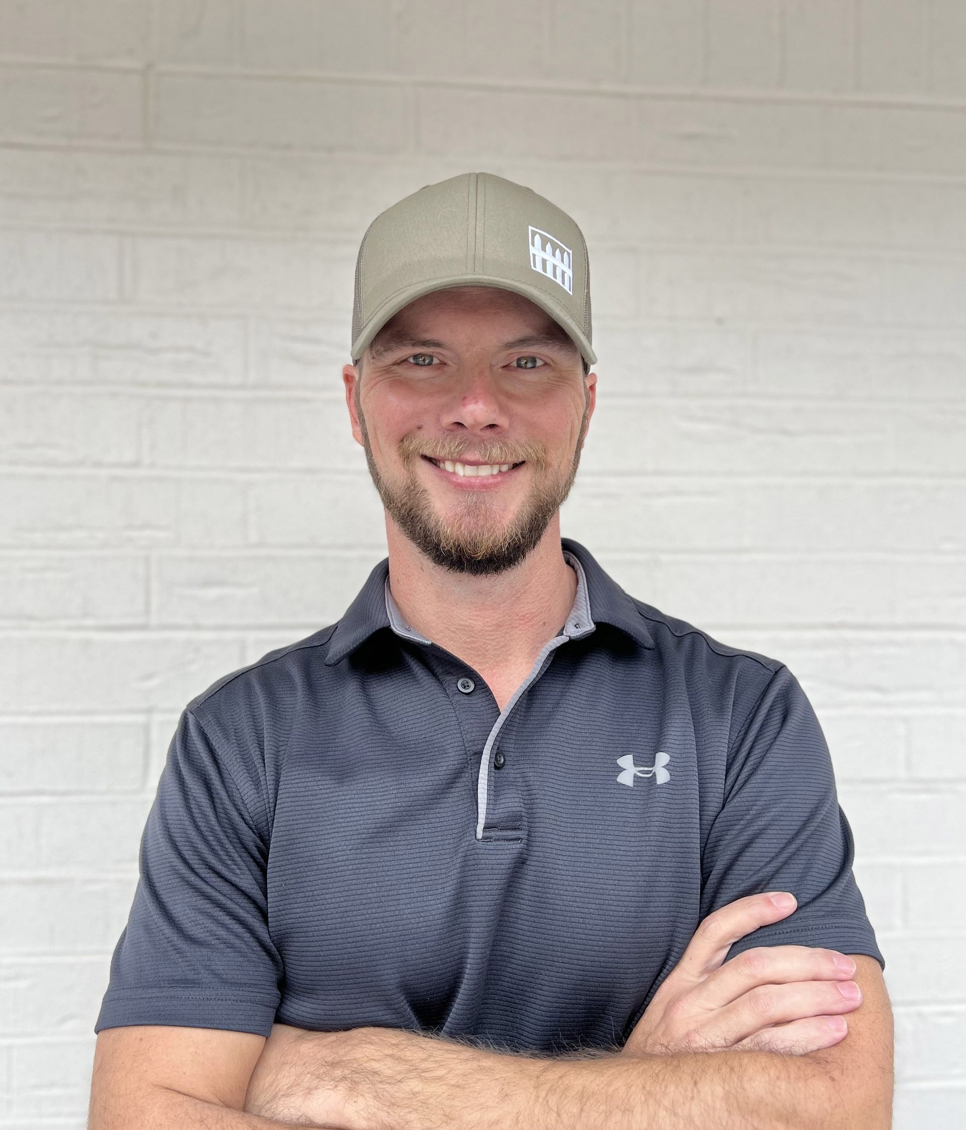 A smiling person with a beard, wearing an olive green baseball cap and a dark patterned Under Armour polo shirt.