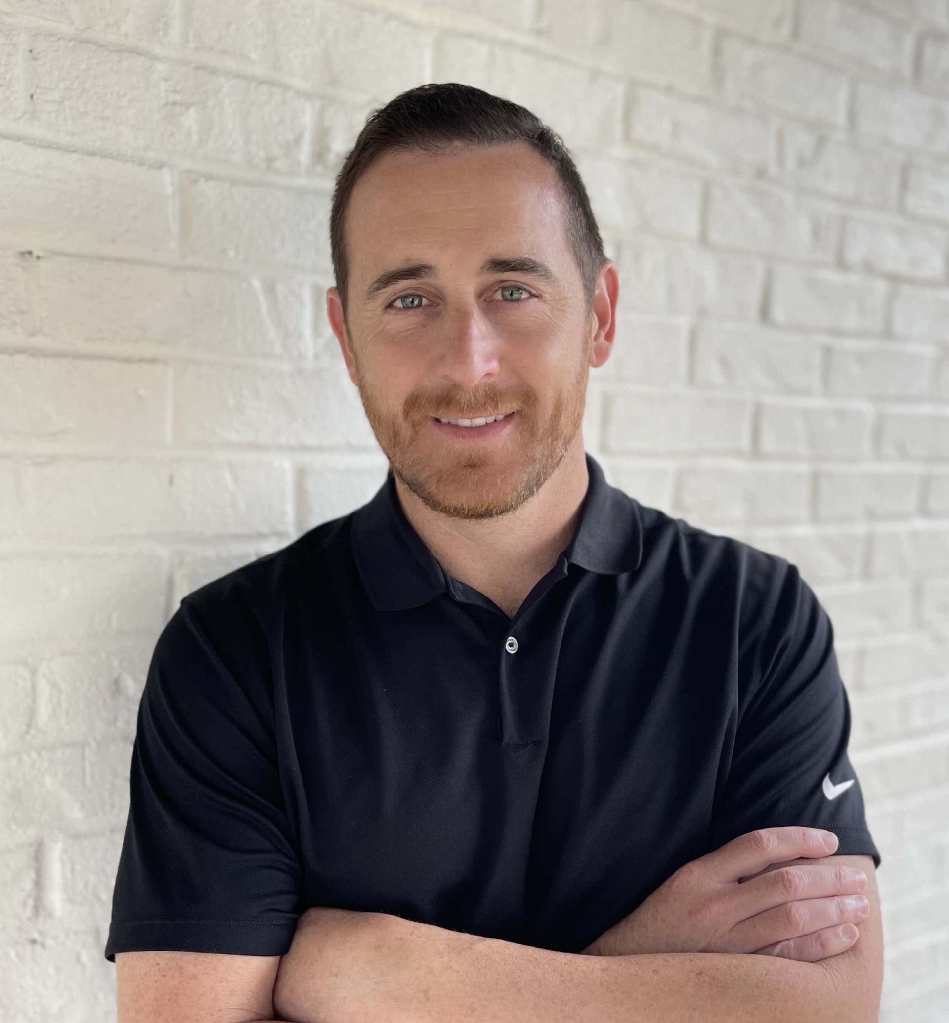 A person with short brown hair and a beard, wearing a black polo shirt, standing with arms crossed against a brick wall.