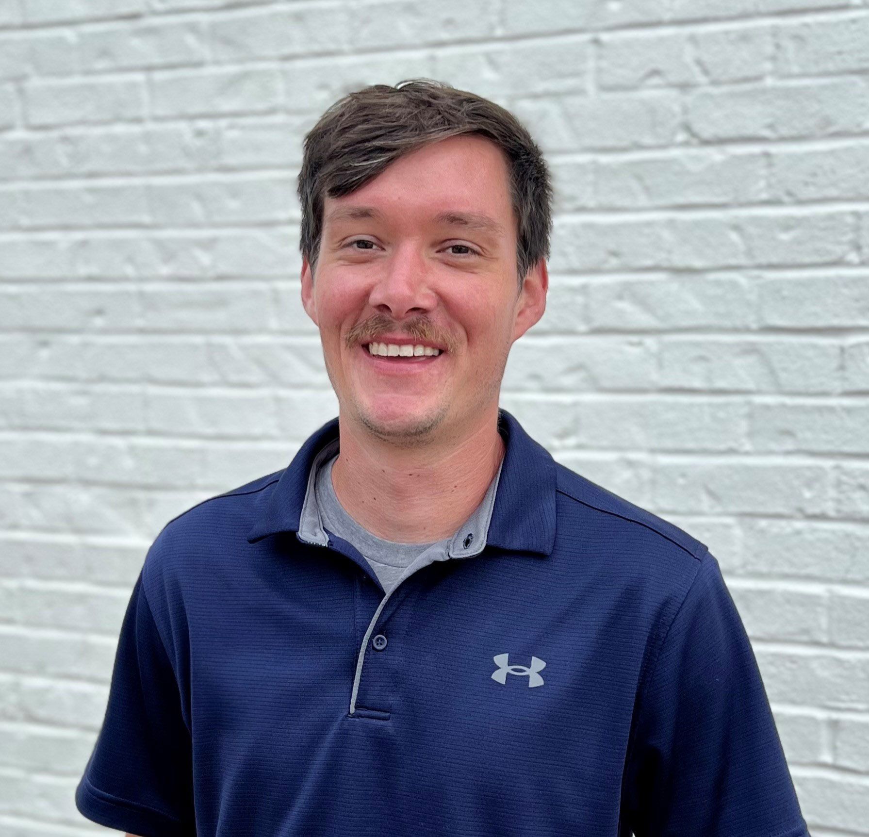 A smiling person with dark hair and a mustache wearing a navy blue polo shirt, standing in front of a white brick wall.