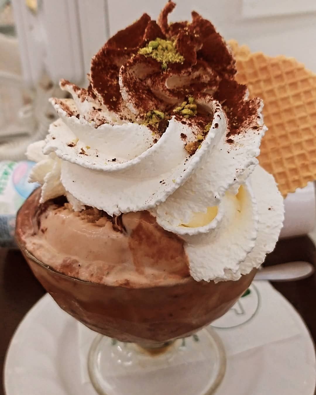 Coppa gelato al cioccolato in bicchiere, guarnita con panna montata, cacao in polvere, pistacchio e waffle.