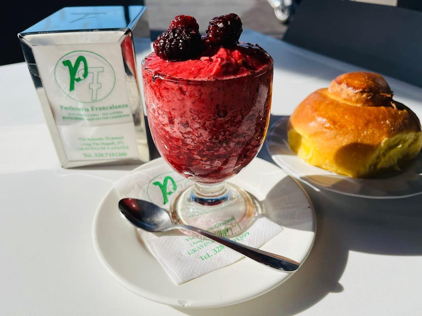 Un bicchiere di granita ai frutti di bosco con more, un cucchiaio, una brioche e un tovagliolo su un tavolo.