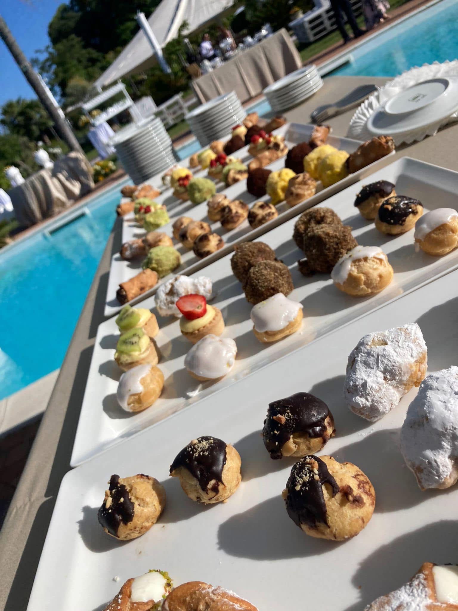 Dessert disposti su vassoi bianchi a bordo piscina; tra i dolci figurano cannoli, dolcetti glassati e ricoperti di cioccolato.