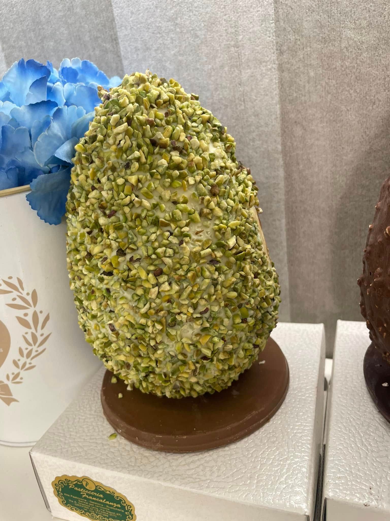 Uovo di Pasqua al cioccolato ricoperto di pistacchi tritati, adagiato su una base di cioccolato fondente.