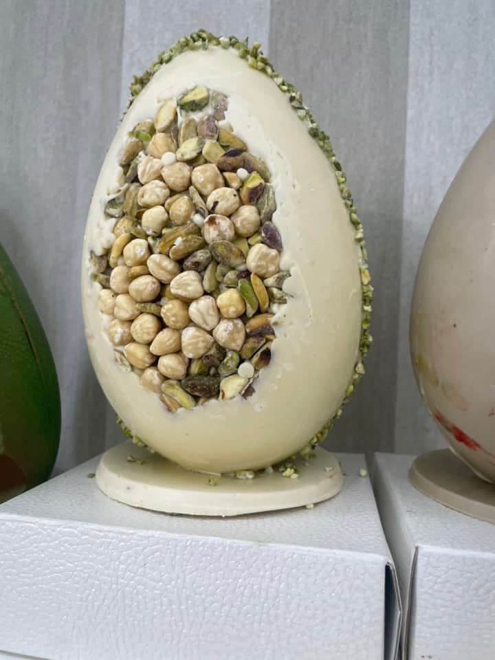 Uovo di Pasqua di cioccolato bianco ripieno di pistacchi e noci, esposto su un espositore bianco.