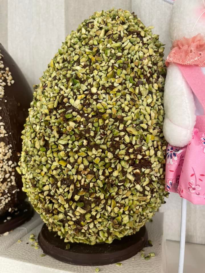 Uovo di cioccolato ricoperto di pezzetti di pistacchio verde, appoggiato su una base di cioccolato fondente, accanto a una figura di coniglietto bianco.
