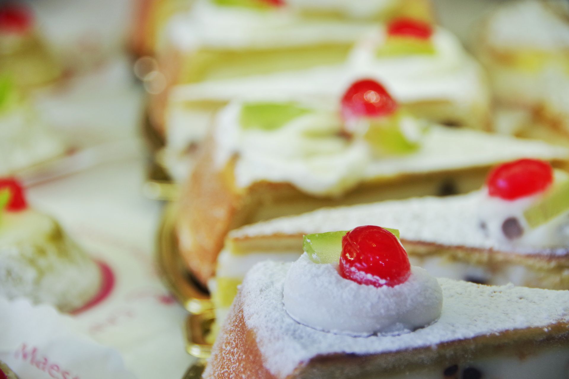 Fette di torta guarnite con panna montata, ciliegie rosse e kiwi verdi, esposte su un vassoio.