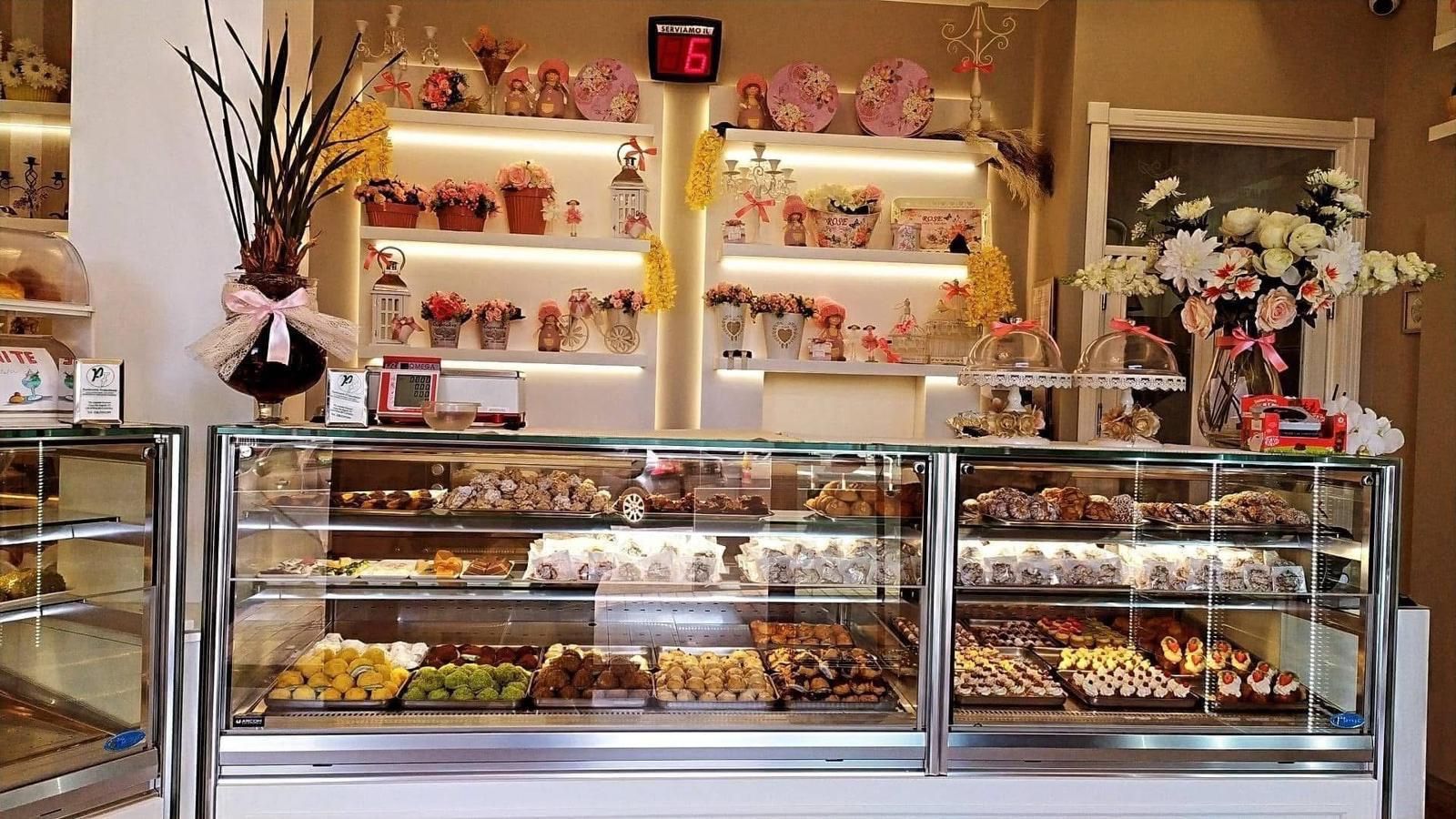 Vetrina da panetteria piena di pasticcini; scaffali sopra con composizioni floreali e regali.