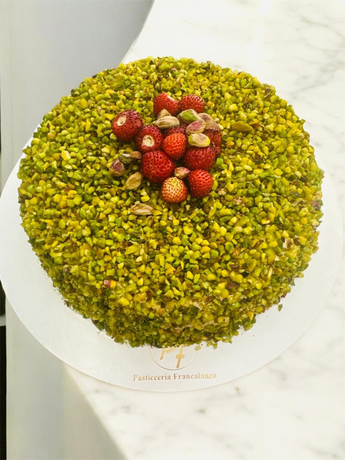 Torta ricoperta di crumble di pistacchi verdi, guarnita con fragole e pistacchi, su un piatto bianco.