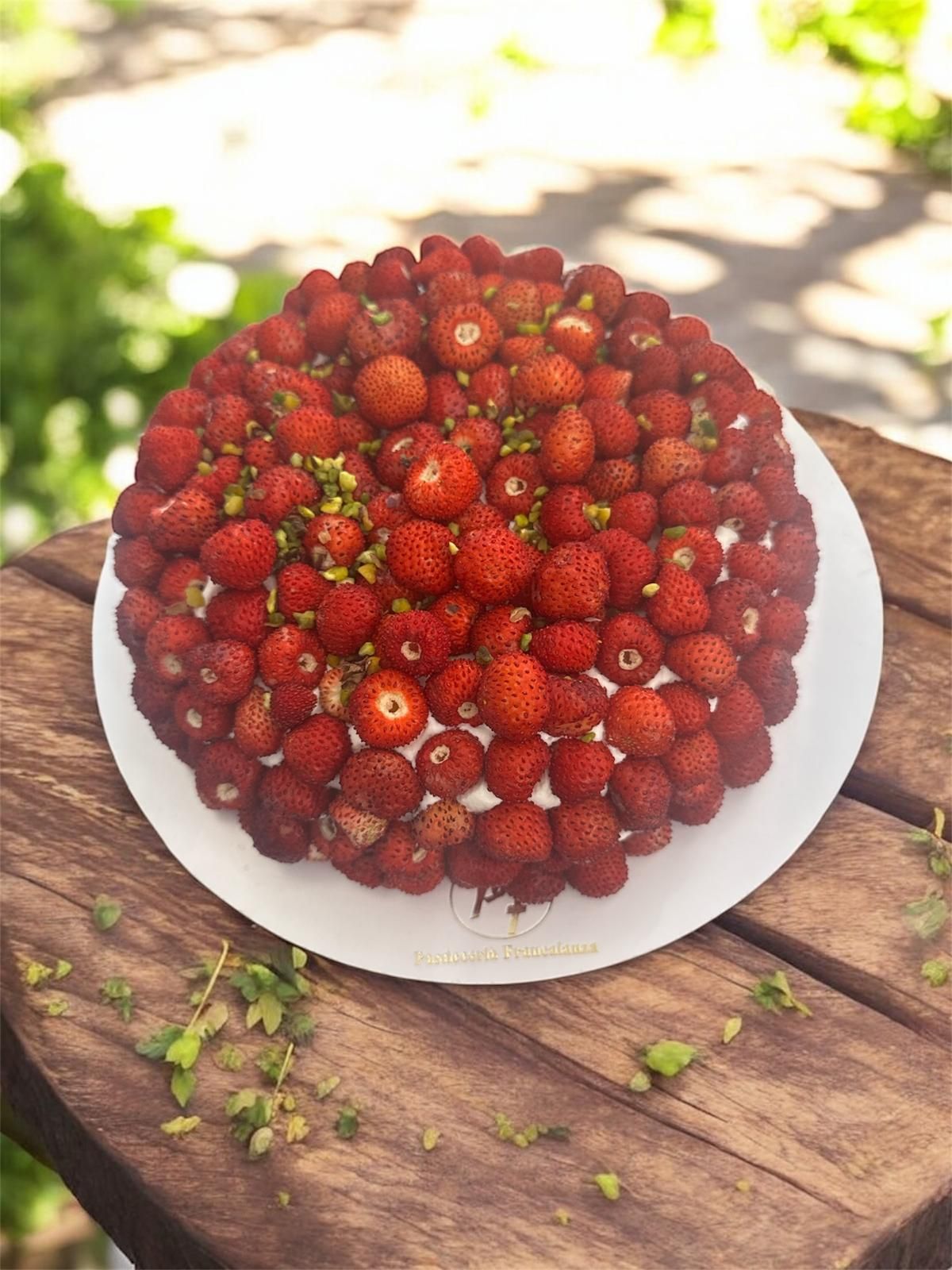 Torta ricoperta di fragole su un piatto bianco, posta su un tavolo di legno, all'aperto con del verde sullo sfondo.