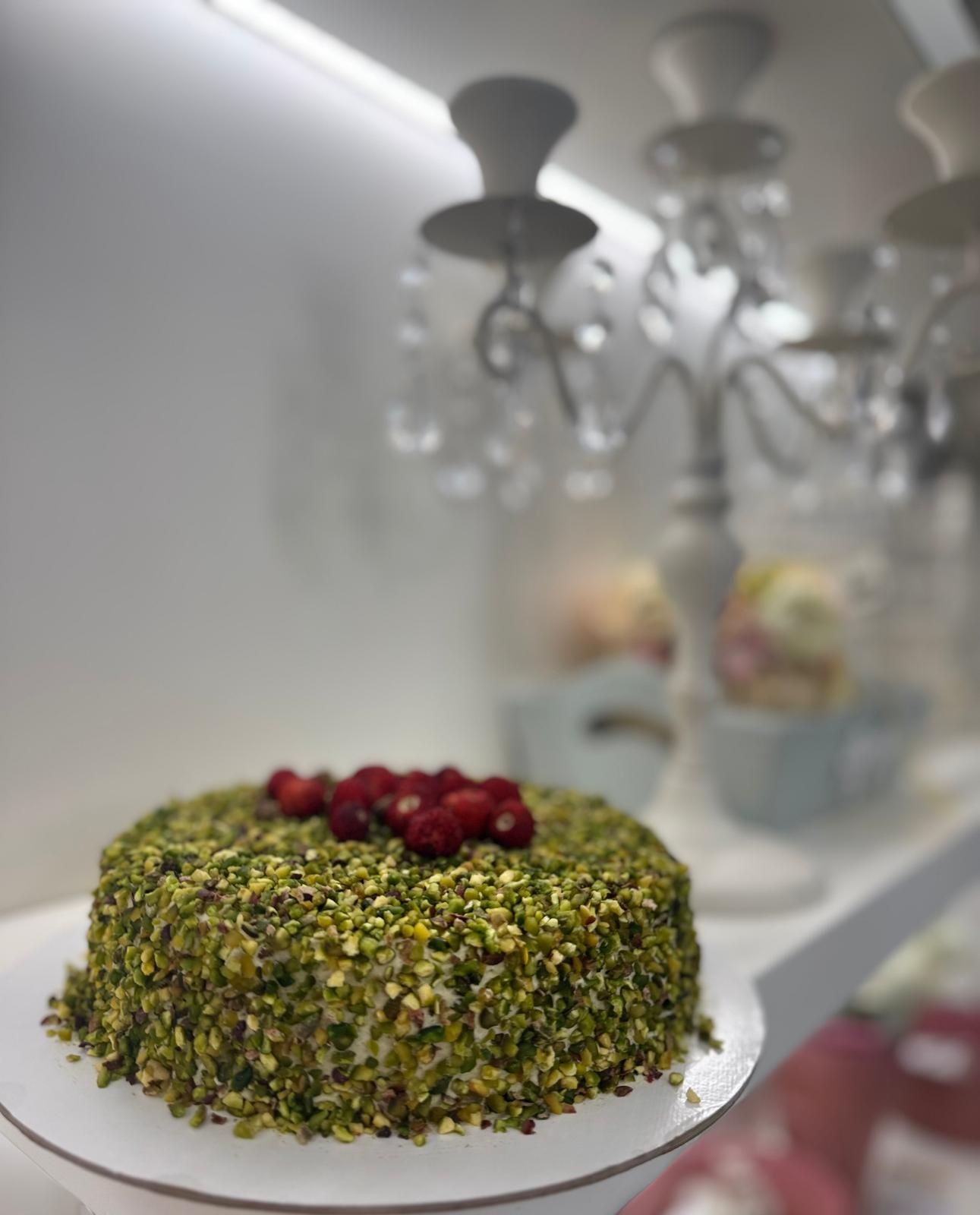 Torta ricoperta di pistacchi e guarnita con frutti di bosco rossi su un'alzata bianca, sfondo sfocato.