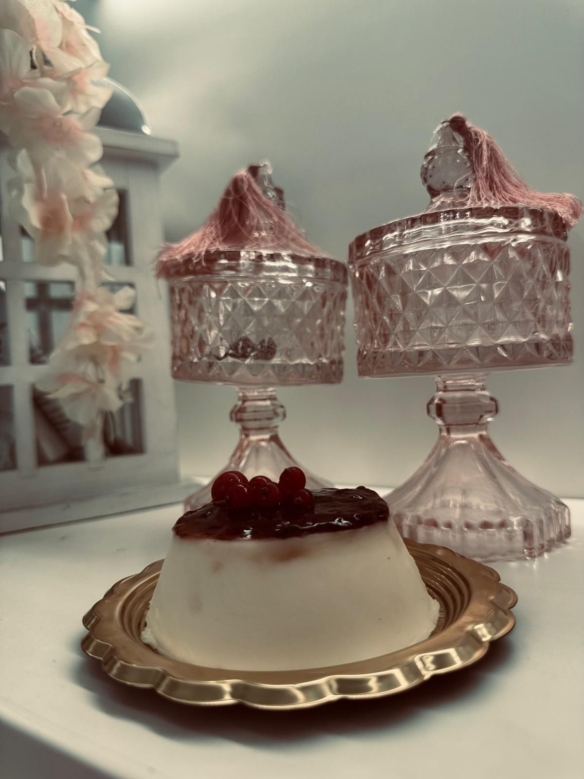 Dessert di panna cotta con frutti rossi su un piatto dorato, con barattoli di vetro rosa su piedistalli sullo sfondo.