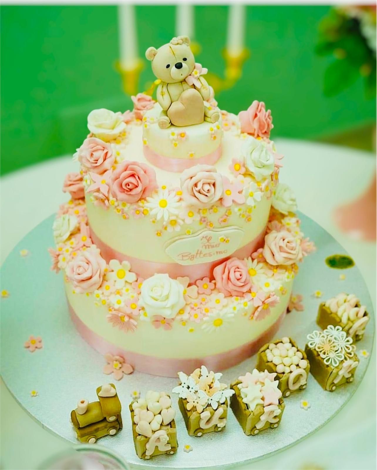Torta a due piani con rose rosa, margherite e un orsacchiotto in cima, circondata da piccoli dolcetti decorati.