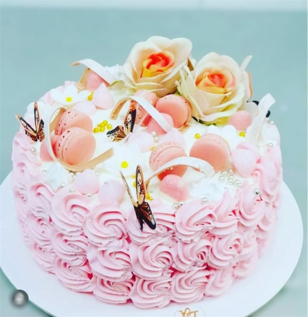 Torta glassata rosa con rose, macaron e farfalle su un piatto bianco.