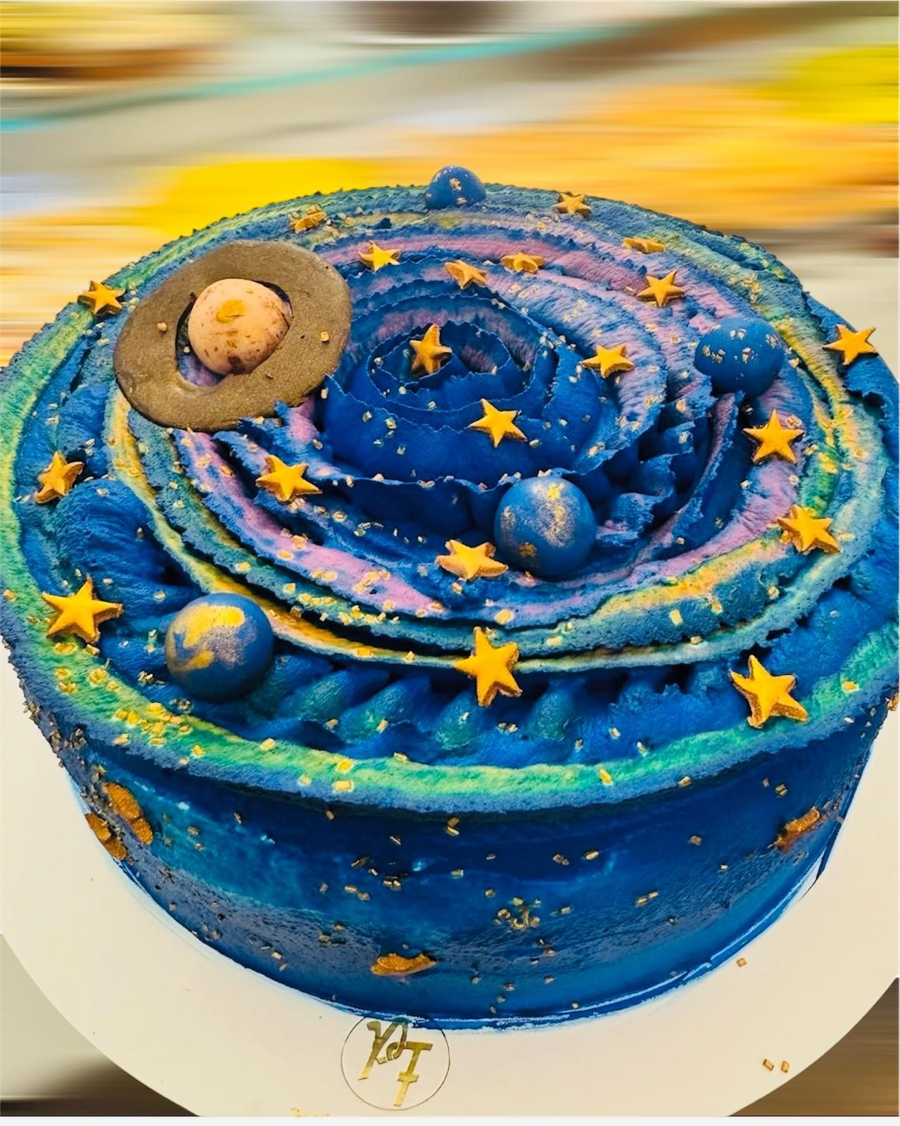 Torta a tema galassia con glassa blu, viola e oro, decorata con stelle, pianeti e un pianeta con anelli.